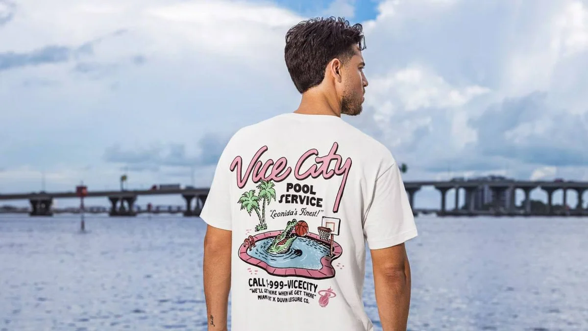 Back of a white Vice City Pool Service tee with an alligator holding a basketball in a pool, the lines Leonida's Finest!, Call 1-999-VICECITY and Miami Heat x Duvin Leisure Co., worn by a man by the water in Miami (Miami Heat store photo)