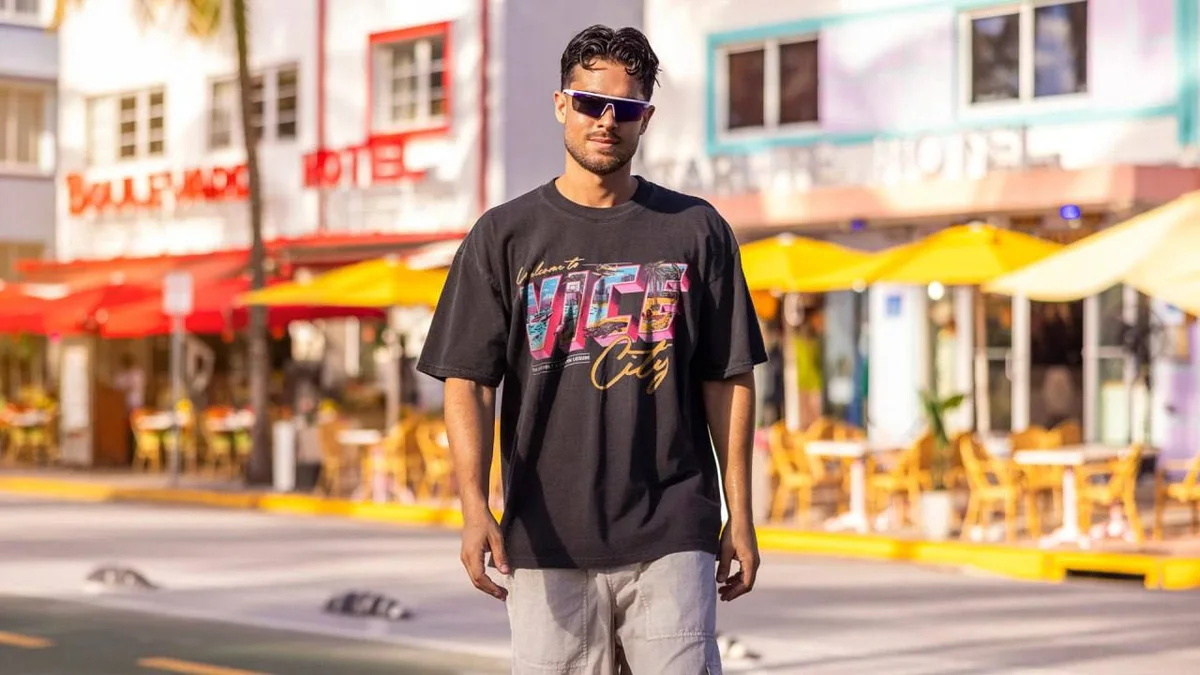 Man on a Miami Beach street lined with hotel signs, wearing the black Court Culture x Duvin 'Vice City Postcard' tee with a postcard-style Welcome to Vice City print (Miami Heat store photo)