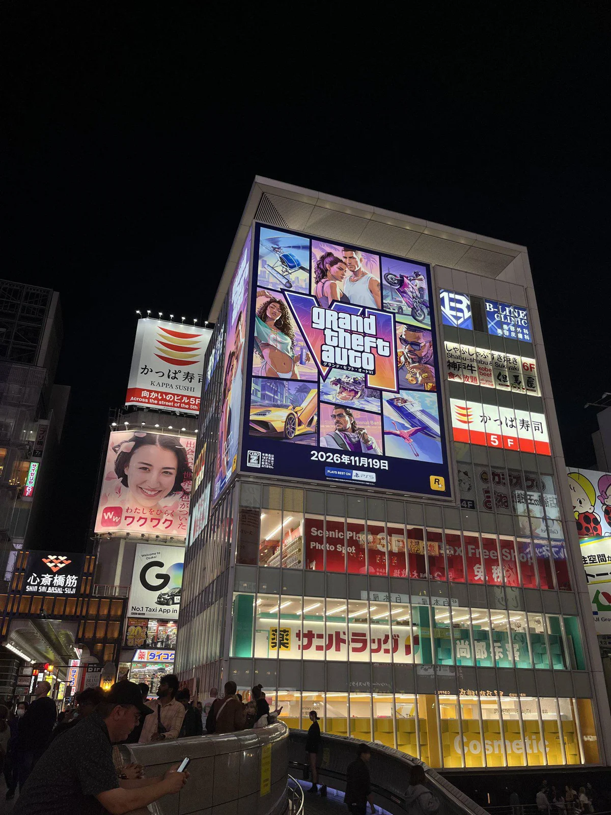 Street view of the GTA 6 billboard in Osaka at night, a collage of official artwork with the logo and 2026年11月19日, people walking below